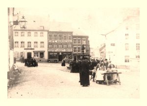 De gauche à droite, dans la rangée d'immeubles bordant la place du Marché : un premier commerce, propriété de Victoire Dehanne en 1912, devenu magasin Delaisse, puis Shoe Post, et aujourd'hui Office du Tourisme; le Café Marischal, aussi épicerie, vendu en 1926, abrita la Maison du Peuple et, depuis 2012, une pizzeria; un magasin A. Petit, maintenant restaurant;  lhôtel Petit, en attente de réaffectation.